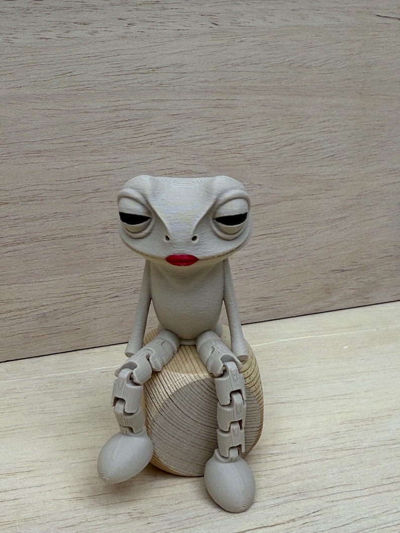May include: A whimsical, gray frog figurine with articulated limbs and a bored expression. The frog is seated on a wooden, rounded base and has red lipstick. The frog's design is reminiscent of a puppet or toy.