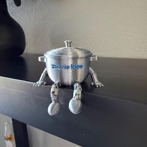 May include: A silver-colored, 3D-printed pot with arms and legs, and the words "Puerto Rico" in blue. The pot is sitting on a black surface. The pot has a lid and handles.