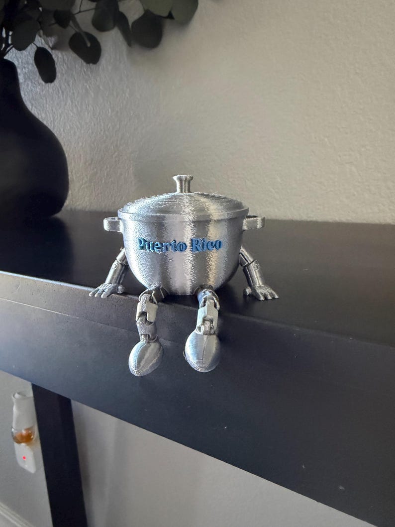 May include: A silver-colored, 3D-printed pot with arms and legs, and the words "Puerto Rico" in blue. The pot is sitting on a black surface. The pot has a lid and handles.