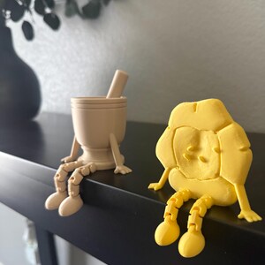 May include: Two whimsical figurines sit on a black surface. One is a tan mortar and pestle with articulated legs. The other is a yellow, cartoonish figure with jointed legs. Both are designed to sit on a shelf or ledge.