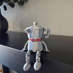 May include: A silver-colored, articulated coffee pot figurine with the text "Puerto Rico" on a red band. The figurine has legs and arms, and is sitting on a black surface. A black vase and greenery are in the background.