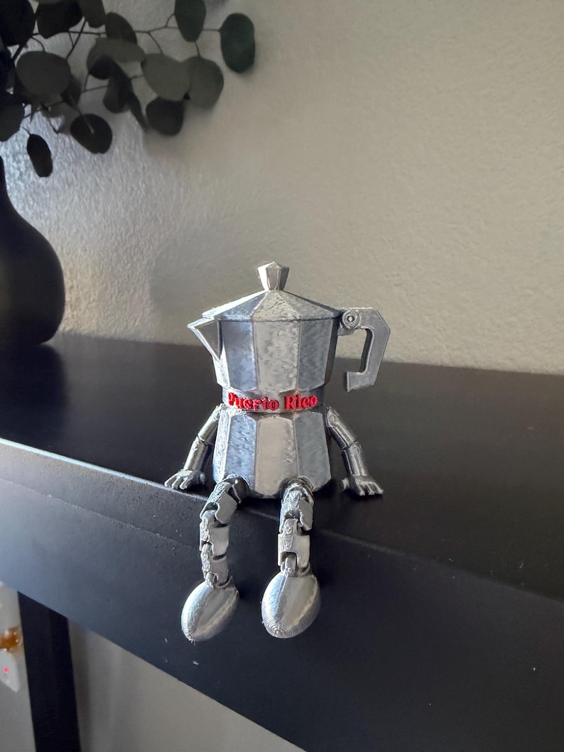 May include: A silver-colored, articulated coffee pot figurine with the text "Puerto Rico" on a red band. The figurine has legs and arms, and is sitting on a black surface. A black vase and greenery are in the background.