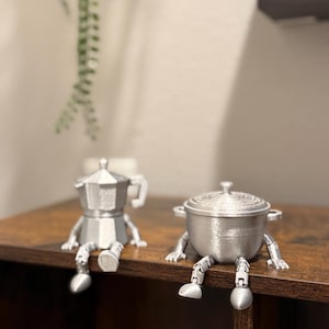 May include: Two silver-colored, 3D-printed kitchen-themed figurines sit on a dark brown surface. One is a coffee maker, the other a pot, both with attached legs and feet. The figurines are designed to sit on the edge of a shelf.