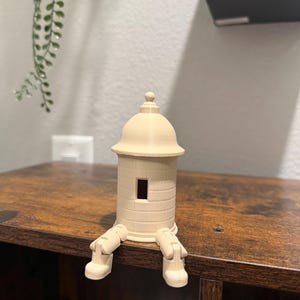 May include: A beige 3D-printed decorative figurine of a small tower with a domed roof and rectangular window. The tower has attached legs, giving it a whimsical appearance. The figurine is sitting on a wooden surface.