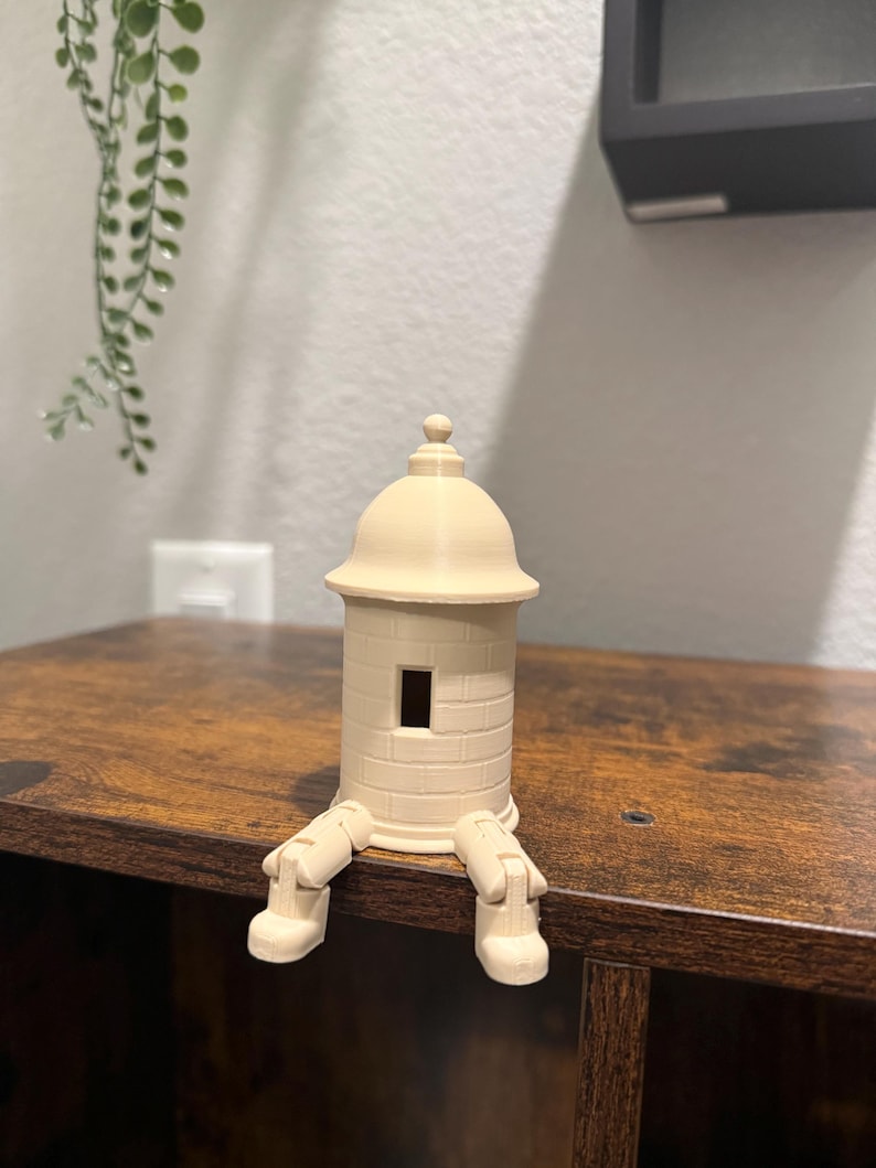 May include: A beige 3D-printed decorative figurine of a small tower with a domed roof and rectangular window. The tower has attached legs, giving it a whimsical appearance. The figurine is sitting on a wooden surface.