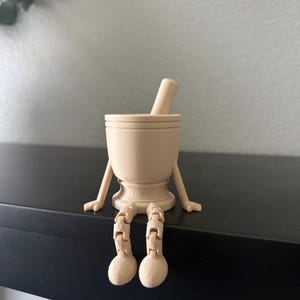 May include: A beige mortar and pestle with arms and legs, sitting on a black shelf. The mortar has a rounded base and a cylindrical shape, with the pestle resting inside. The arms and legs are articulated, giving the mortar a whimsical appearance.