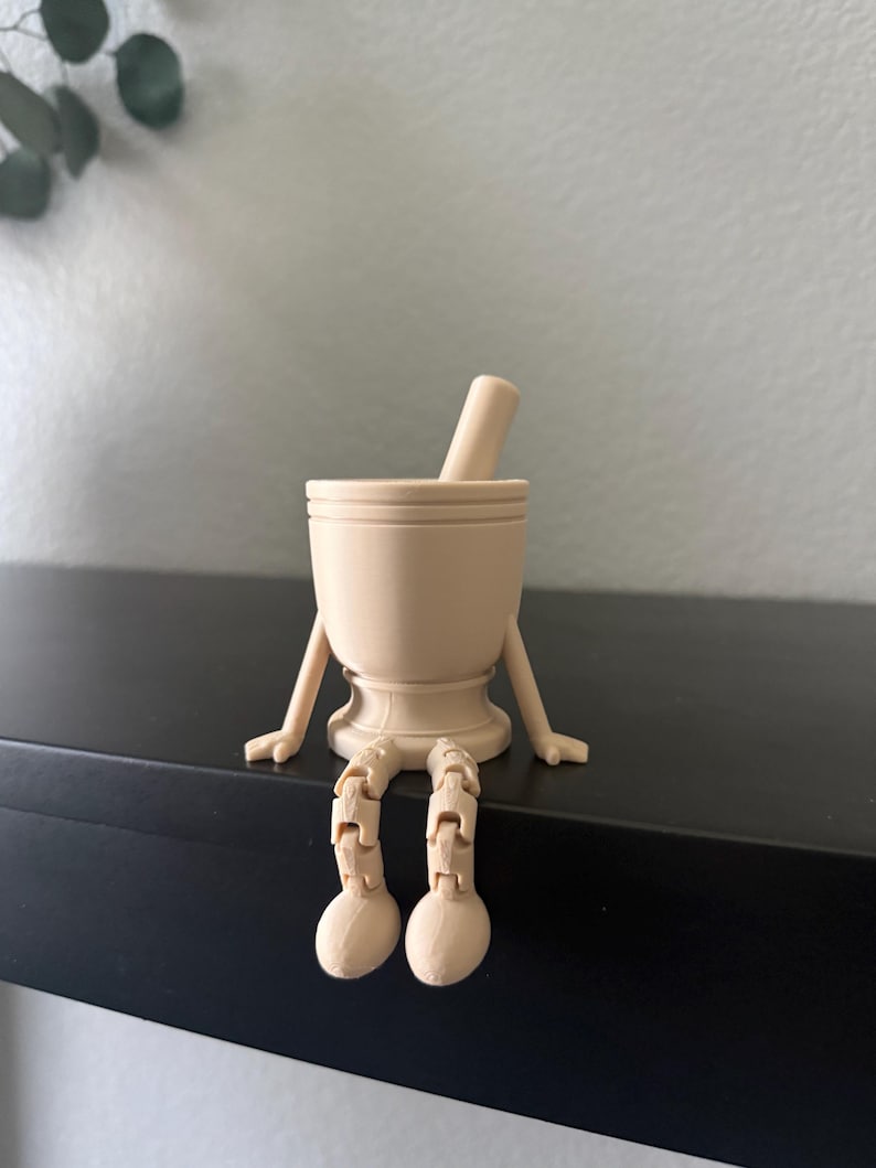 May include: A beige mortar and pestle with arms and legs, sitting on a black shelf. The mortar has a rounded base and a cylindrical shape, with the pestle resting inside. The arms and legs are articulated, giving the mortar a whimsical appearance.