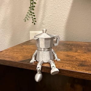 May include: A silver-colored, 3D-printed coffee maker figurine with arms and legs, sitting on a wooden surface. The figurine is a whimsical representation of a coffee pot with a handle and a lid. The background includes a wall and a hanging plant.