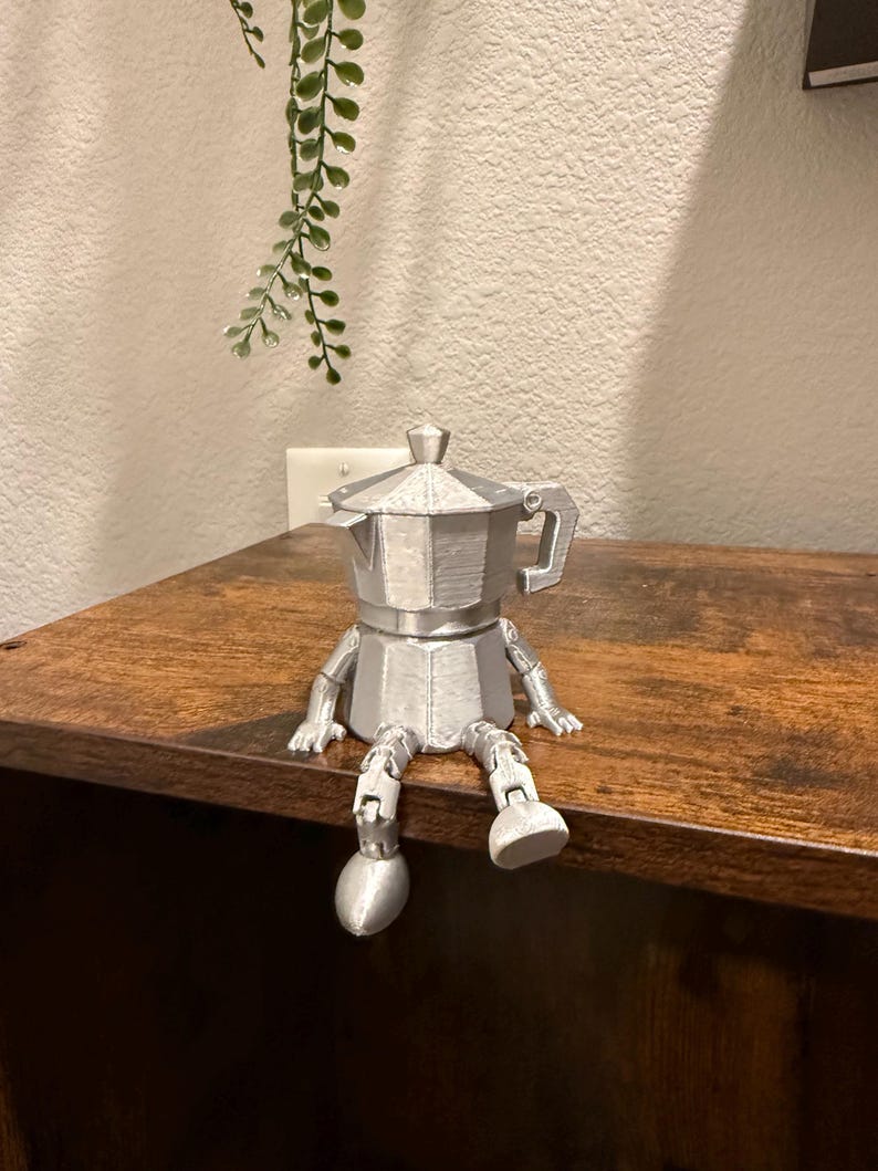 May include: A silver-colored, 3D-printed coffee maker figurine with arms and legs, sitting on a wooden surface. The figurine is a whimsical representation of a coffee pot with a handle and a lid. The background includes a wall and a hanging plant.