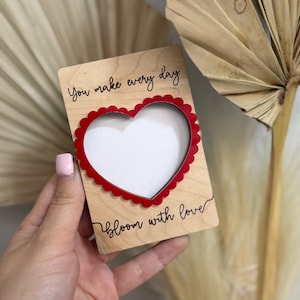 Puede incluir: Una tarjeta de madera con un marco en forma de corazón rojo y las palabras "You make every day" y "bloom with love" en escritura negra. La tarjeta se sostiene contra un fondo neutro.