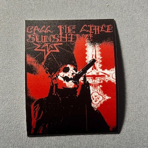 May include: A rectangular sticker with a black background and red accents. The sticker features a stylized image of a figure in a mask holding a gun, with the text "CALL ME LITTLE SUNSHINE" in red. A red cross is visible.
