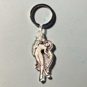 Puede incluir: Un llavero con una ilustración de un ángel desnudo. El ángel tiene grandes alas, un halo y un acento rojo. El diseño es estilizado. El llavero tiene un anillo de metal plateado y un accesorio de plástico transparente.