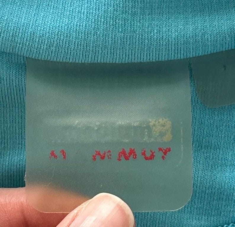 K&ouml;nnte beinhalten: Ein helles t&uuml;rkisfarbenes Kleidungsst&uuml;ck mit einem halbtransparenten, rechteckigen Etikett am Halsausschnitt. Das Etikett zeigt undeutlichen, teilweise verdeckten Text in einer dunklen Farbe und den roten Aufdruck M MUY am unteren Rand.