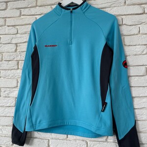 K&ouml;nnte beinhalten: Ein hellblauer Langarm-Pullover mit Viertelrei&szlig;verschluss, schwarzen Seiteneins&auml;tzen und schwarzen &Auml;rmelb&uuml;ndchen. Der Markenname Mammut ist in Rot auf der Brust gestickt, mit einem roten runden Logo am linken &Auml;rmel und einem Polartec-Etikett an der Seite.