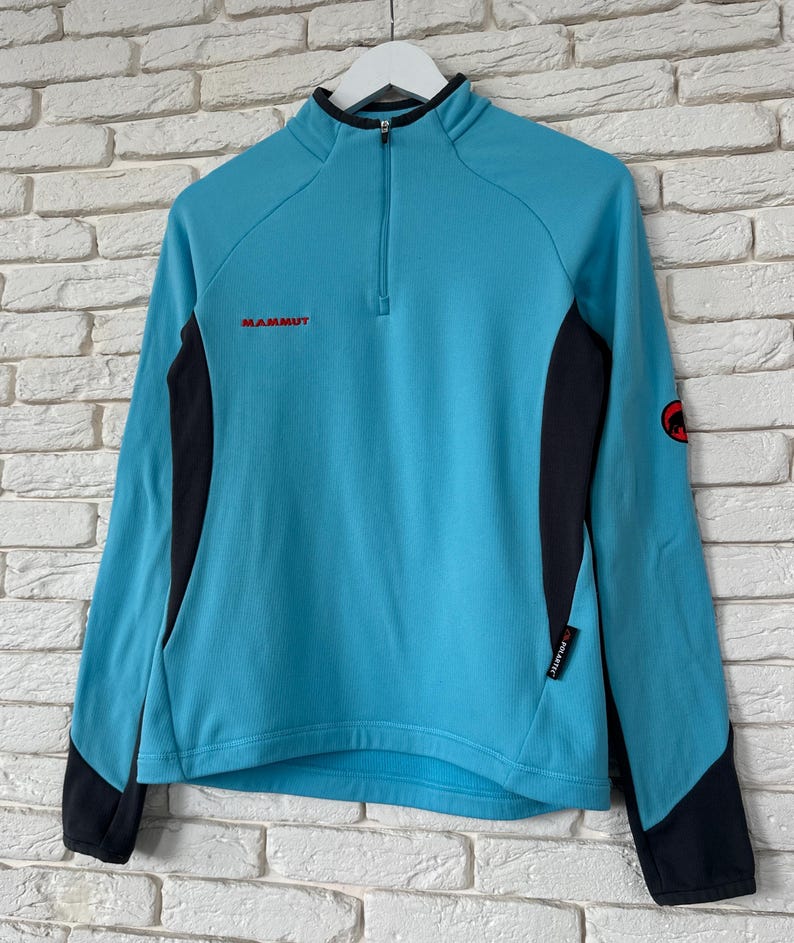 K&ouml;nnte beinhalten: Ein hellblauer Langarm-Pullover mit Viertelrei&szlig;verschluss, schwarzen Seiteneins&auml;tzen und schwarzen &Auml;rmelb&uuml;ndchen. Der Markenname Mammut ist in Rot auf der Brust gestickt, mit einem roten runden Logo am linken &Auml;rmel und einem Polartec-Etikett an der Seite.