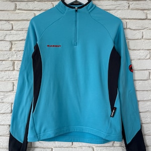 K&ouml;nnte beinhalten: Ein hellblauer Langarm-Pullover mit schwarzem Viertelrei&szlig;verschluss am Kragen und schwarzen Seiteneins&auml;tzen. Das Mammut-Logo ist in Rot auf der Brust gestickt, mit einem kleinen schwarzen Polartec-Etikett an der Seite und einem runden Mammut-Logo am linken &Auml;rmel.