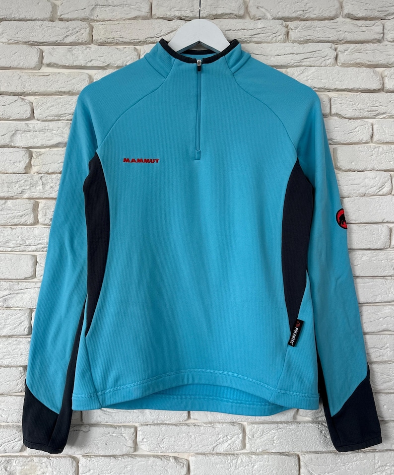 K&ouml;nnte beinhalten: Ein hellblauer Langarm-Pullover mit schwarzem Viertelrei&szlig;verschluss am Kragen und schwarzen Seiteneins&auml;tzen. Das Mammut-Logo ist in Rot auf der Brust gestickt, mit einem kleinen schwarzen Polartec-Etikett an der Seite und einem runden Mammut-Logo am linken &Auml;rmel.