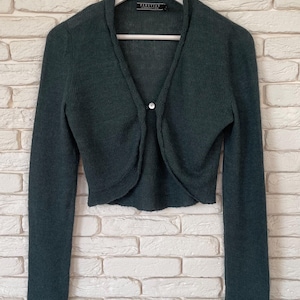 Op de afbeelding: Een donker bosgroen bolero-vest met lange mouwen en een enkele knoopsluiting aan de voorzijde. Het gebreide kledingstuk heeft een ronde halslijn en uitlopende boorden aan het uiteinde van elke mouw. Een label met Peruvian Connection is zichtbaar.