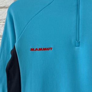 K&ouml;nnte beinhalten: Ein hellblauer Langarm-Pullover mit einem viertellangen Rei&szlig;verschluss am Kragen und schwarzen Seiteneins&auml;tzen. Der Markenname MAMMUT ist in rotem Garn auf der linken Brustseite des strukturierten Kleidungsst&uuml;cks eingestickt.