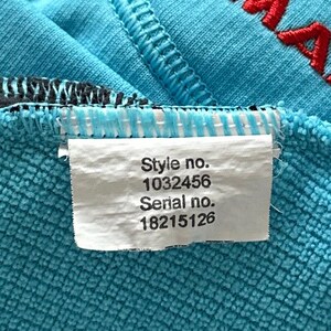 K&ouml;nnte beinhalten: Ein leuchtend t&uuml;rkisfarbener Stoff mit strukturierter Oberfl&auml;che, roter Stickerei und einem wei&szlig;en rechteckigen Etikett. Das Etikett zeigt den Text Style no. 1032456 und Serial no. 18215126 in schwarzer Schrift auf dem t&uuml;rkisen Material.