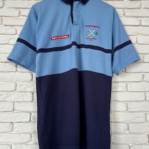 May include: A two-tone rugby shirt in light blue and navy, with short sleeves and a navy collar. The shirt features a three-button placket. The text "BAE SYSTEMS" is printed on the chest, and "LEEUWENS NOMADS RC SOUTH AFRICA 1993" is on the right.