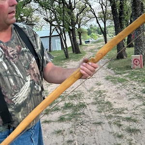 Puede incluir: Un arco de madera artesanal, sostenido por una persona. El arco es de color marrón claro y tiene una cuerda. El fondo incluye árboles, un camino de tierra y una diana.