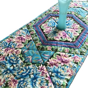 May include: A blue and green table runner with a floral pattern and dragonfly accents. The runner is quilted and has a decorative border. A blue glass vase is placed in the centre of the runner.