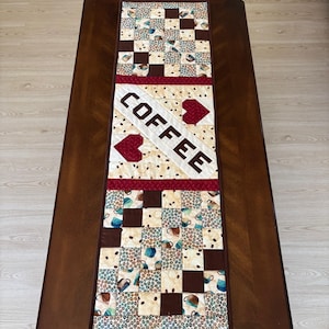May include: A quilted coffee table runner with a patchwork design. The runner features squares of coffee cups, coffee beans, and solid brown squares. The word "COFFEE" is displayed in large white letters with red heart accents.