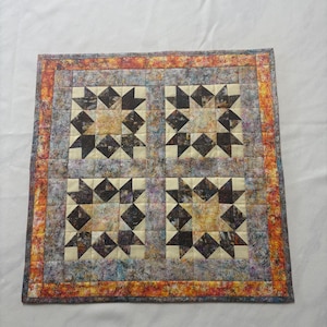 Peut inclure: Une pièce textile matelassée carrée avec quatre motifs d'étoiles. Chaque étoile est composée de tissu marron foncé et crème. La bordure présente des teintes orange, grises et bleues. Le fond est blanc.