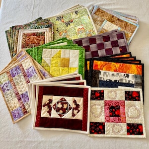 May include: A collection of rectangular fabric placemats featuring various quilted patterns, including floral, geometric, heart, and elephant motifs in diverse colours such as red, yellow, green, purple, black, and cream, arranged in a layered stack.