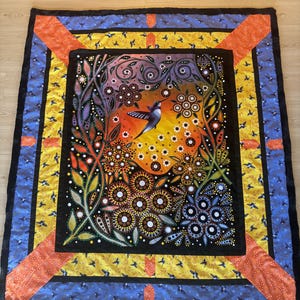 May include: A colourful quilt with a black background featuring a hummingbird in flight surrounded by flowers and leaves. The quilt has a multi-coloured border with a repeating pattern of small birds.