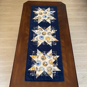May include: A rectangular quilted table runner featuring three large star-shaped appliques. The stars are made of a cream-coloured fabric printed with yellow suns and blue moons, set against a dark navy blue background with white star constellations.