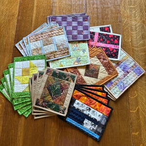 May include: A collection of rectangular fabric coasters featuring various quilted patterns, including floral, geometric, and animal prints in colours such as green, yellow, brown, purple, black, and pink, arranged in a layered, overlapping display.