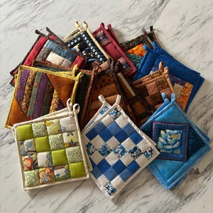 May include: A collection of square fabric potholders featuring various patchwork designs, quilted textures, and patterns including floral prints, checkered motifs, and solid colours in shades of green, blue, brown, red, and yellow with hanging loops.