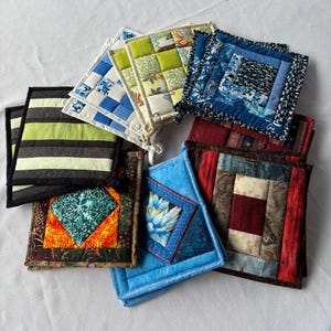 May include: A collection of square quilted potholders featuring various fabric patterns including green and black stripes, blue floral patchwork, red and brown geometric blocks, and a bright orange and teal square with a floral centre design.