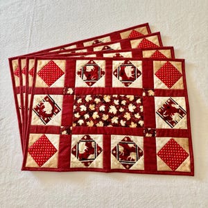 May include: A set of four rectangular quilted placemats featuring a patchwork design with red and cream fabric. The pattern includes diamond-shaped blocks with buffalo silhouettes, red fabric with white dots, and a central panel with maple leaves.
