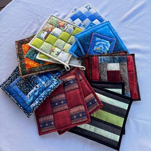 May include: A collection of square fabric pot holders featuring various patchwork designs, including floral patterns, geometric stripes, and solid colours in shades of blue, green, red, and brown, arranged in a layered, overlapping circular formation.