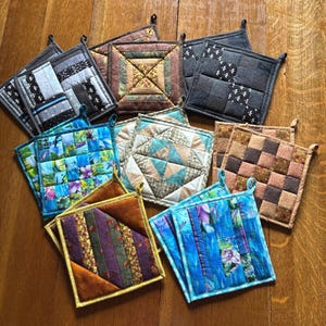 Peut inclure: Une collection de maniques carrées matelassées présentant divers motifs de tissus, notamment des motifs floraux bleus, des carreaux marron, des formes géométriques grises et des rayures multicolores. Chaque carré possède une petite boucle.