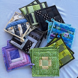 May include: A collection of square quilted potholders in various colours and patterns, including shades of green, purple, blue, and black. The designs feature geometric patchwork, floral prints, and celestial motifs with sun and moon illustrations.