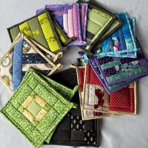 May include: A collection of square quilted pot holders arranged in a circular pattern. The pot holders feature various fabric designs including floral, geometric, and star patterns in colours such as green, purple, blue, red, and black.