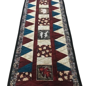May include: A long, rectangular table runner with a patchwork design. The runner features a variety of fabrics in shades of red, brown, blue, and white. The centre of the runner features a large leaf design, while the sides are decorated with smaller leaf and animal motifs.