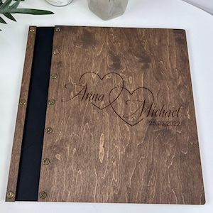 Puede incluir: Libro de visitas de boda de madera teñida oscura. La portada presenta dos corazones entrelazados con los nombres "Anna" y "Michael" y la fecha "25.05.2022" grabados. El lomo es negro y el libro está sujeto con tornillos color latón.