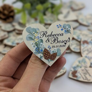 May include: A wooden heart-shaped wedding favor with the names "Rebecca & Bosco" and the date "24.01.2026" printed on it. The design includes floral and pinecone illustrations in blue, yellow, and green. Other similar favors are in the background.