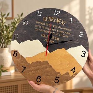 May include: A round wooden wall clock featuring a mountain landscape design in shades of dark gray, cream, and light brown. The clock face displays the text "RETIREMENT," "REMEMBER THE PAST WITH FONDNESS," "LOOK TO THE FUTURE WITH EAGERNESS," "LIVE EVERY MOMENT TO THE FULLEST," "COLOSSIANS 3:11," and "1983-2024" with black numbers 1 through 12.