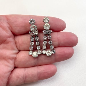 Puede incluir: Un par de pendientes colgantes plateados. Cada pendiente presenta brillantes pedrería transparente de varios tamaños. El diseño incluye un grupo de piedras en la parte superior, una sección central y una piedra más grande en la parte inferior.