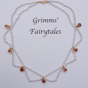 Puede incluir: Un collar de doble cadena de perlas con dijes de manzana roja. El collar tiene un cierre dorado y está decorado con pequeñas perlas blancas. El texto "Grimms' Fairytales" está escrito en negro sobre un fondo blanco.