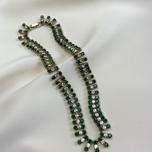 Puede incluir: Un collar dorado con filas alternas de cristales verde esmeralda y transparentes. El collar está colocado sobre una superficie de satén blanco. El collar tiene un cierre de broche y un diseño único.