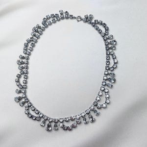 May include: A silver necklace adorned with clear rhinestones. The necklace has a tiered design with multiple rows of stones and teardrop-shaped dangles. The clasp is visible at the back. The jewellery is displayed on a white, silky background.