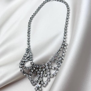 May include: A silver-toned statement necklace with a bib design. The necklace features numerous clear, sparkling rhinestones of various sizes, set in a delicate, ornate pattern. The necklace is displayed on a white satin background.