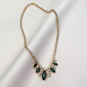 May include: A gold-coloured necklace adorned with clear and emerald green rhinestones. The necklace has a delicate chain and a central pendant design of marquise-cut green stones and smaller clear stones.
